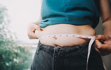 Overweight woman pinching belly fat while holding tape measure around her waist