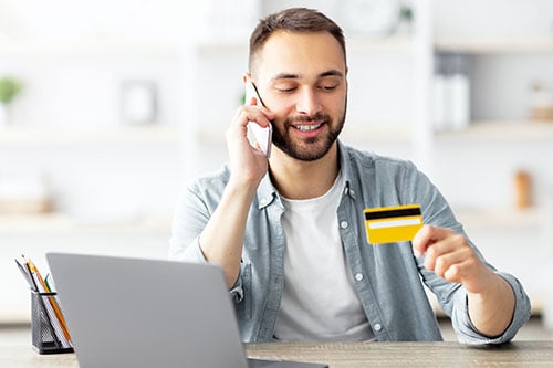 Smiling young man sitting at kitchen table paying his bill with a credit card over the phone